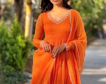 May include: An orange long-sleeved dress with a V-neckline, adorned with delicate white embroidery along the neckline and cuffs. The dress features a flowing skirt and is paired with a matching scarf. The outfit is made of a lightweight fabric.