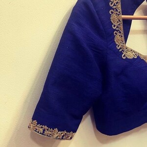 Op de afbeelding: Een donkerblauwe blouse met gouden borduurwerk rond de halslijn en mouwen. De blouse is gemaakt van een zachte, zijdeachtige stof.