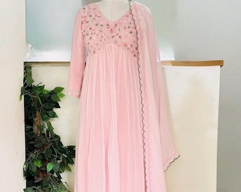 Puede incluir: Un vestido rosa claro con mangas largas y cuello en V. El corpiño está adornado con bordados florales. El vestido tiene una falda fluida y se combina con una bufanda a juego. Los bordes de la bufanda están adornados con un diseño festoneado negro.