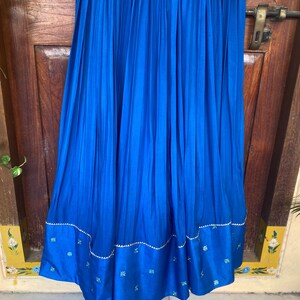 May include: A long, royal blue skirt with pleats and a decorative embroidered trim. The trim features a wavy line of silver sequins and small blue and green floral designs.