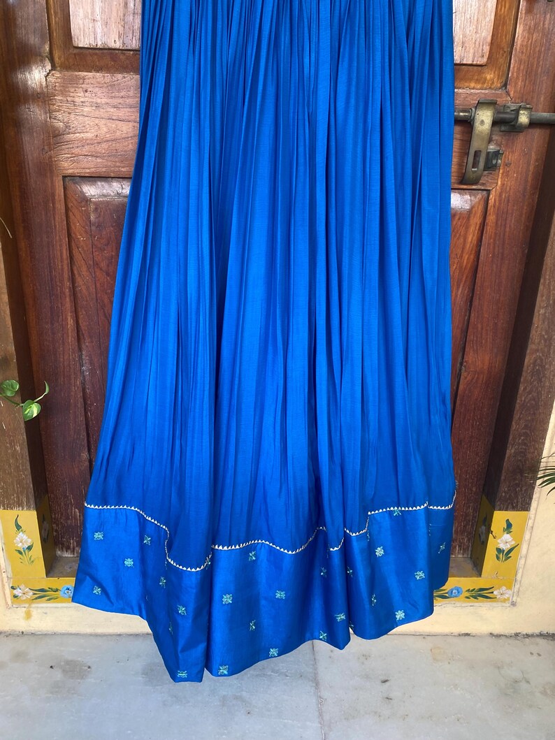 May include: A long, royal blue skirt with pleats and a decorative embroidered trim. The trim features a wavy line of silver sequins and small blue and green floral designs.