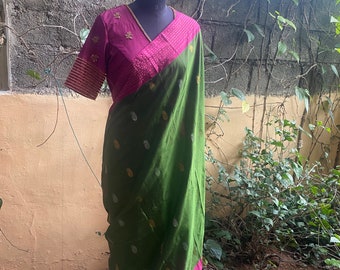 May include: A green and magenta saree displayed on a mannequin. The saree features a green body with gold and white floral patterns. The blouse is magenta with gold embroidery and a V-neck. The sleeves have a striped design.