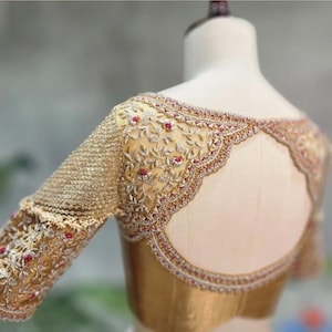 Può includere: Una camicetta dorata, riccamente ricamata, con maniche lunghe. La camicetta presenta dettagliati motivi floreali con accenti rossi e uno scollo smerlato. La schiena ha un design a goccia.