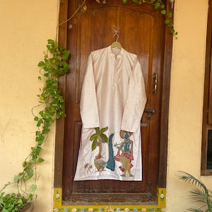 May include: A white kurta with a pink and gold embroidered design featuring a scene of a man playing a flute with a peacock and palm trees. The kurta is hanging on a hanger in front of a wooden door.