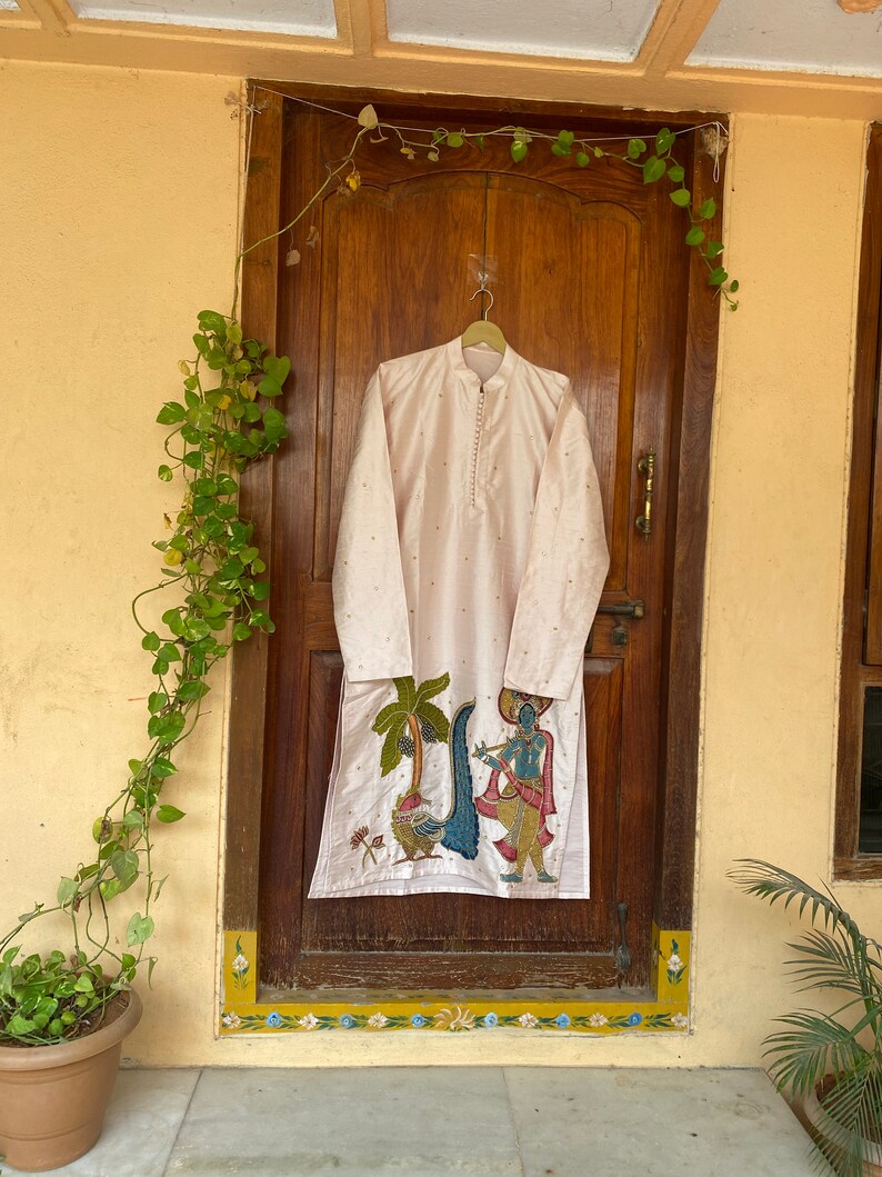 May include: A white kurta with a pink and gold embroidered design featuring a scene of a man playing a flute with a peacock and palm trees. The kurta is hanging on a hanger in front of a wooden door.