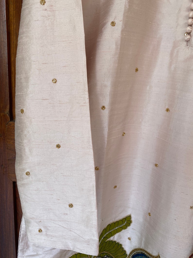 May include: A pale pink garment with gold embellishments. The garment has long sleeves and a decorative design on the bottom.