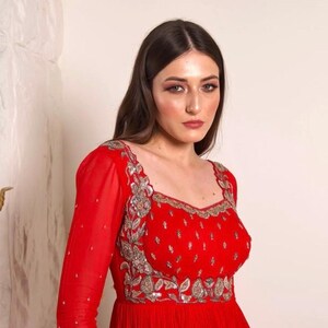 May include: A woman wearing a red dress with silver embroidery. The dress has a square neckline and long sleeves. The woman has long brown hair and is looking at the camera.