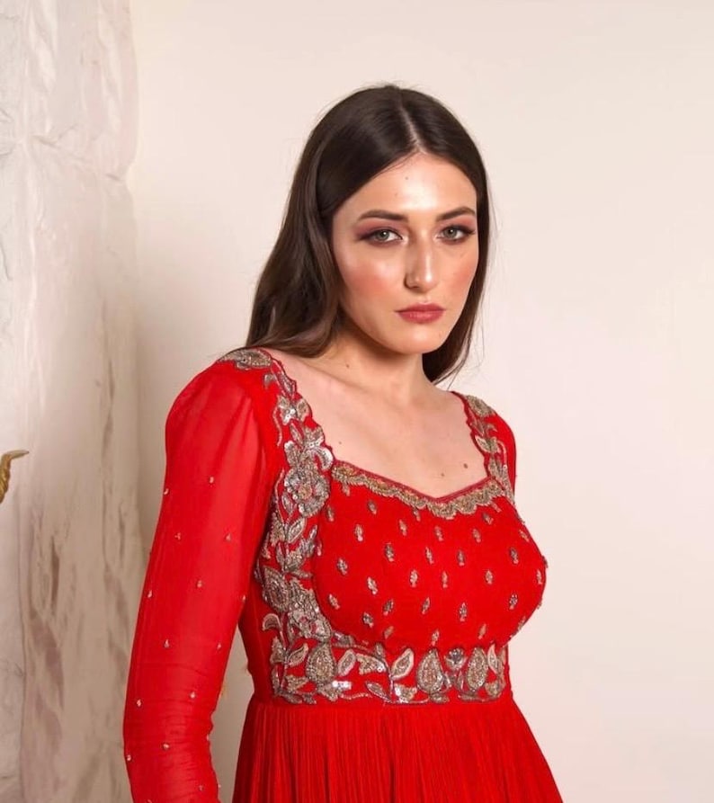 May include: A woman wearing a red dress with silver embroidery. The dress has a square neckline and long sleeves. The woman has long brown hair and is looking at the camera.