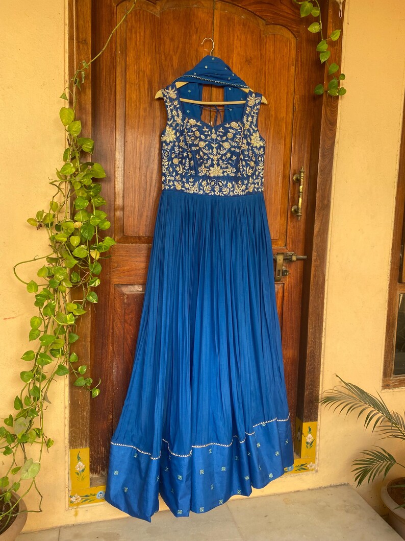 May include: A long, royal blue dress with gold embroidery and a matching dupatta. The dress has a sleeveless bodice and a flowing skirt.