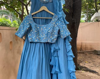 May include: A light blue formal outfit, including a top with embroidered floral details and a matching skirt. The skirt has a pleated design, and the outfit is displayed on a wooden hanger. A matching scarf with ruffled edges completes the ensemble.