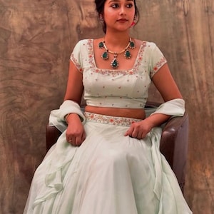 May include: A woman wearing a light green, embroidered, two-piece outfit with a matching dupatta. She is wearing a green and gold necklace with a teardrop design.