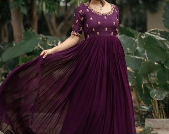 Puede incluir: Una mujer con un vestido largo de color morado fluido con bordados dorados. El vestido tiene una falda plisada y un escote redondo. Está de pie en un jardín.