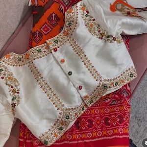 Puede incluir: Blusa color marfil con intrincados bordados en dorado, naranja, verde y rojo. La blusa presenta un diseño detallado de pájaro en la manga y botones coloridos. Se muestra sobre una tela estampada roja y naranja.
