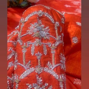 K&ouml;nnte beinhalten: Nahaufnahme eines leuchtend orangefarbenen Kleidungsst&uuml;cks mit aufwendiger Silber- und Goldstickerei. Die detaillierten floralen und geometrischen Muster erzeugen eine luxuri&ouml;se Textur. Der Stoff scheint einen subtilen Glanz zu haben, der den Reichtum des Designs verst&auml;rkt.