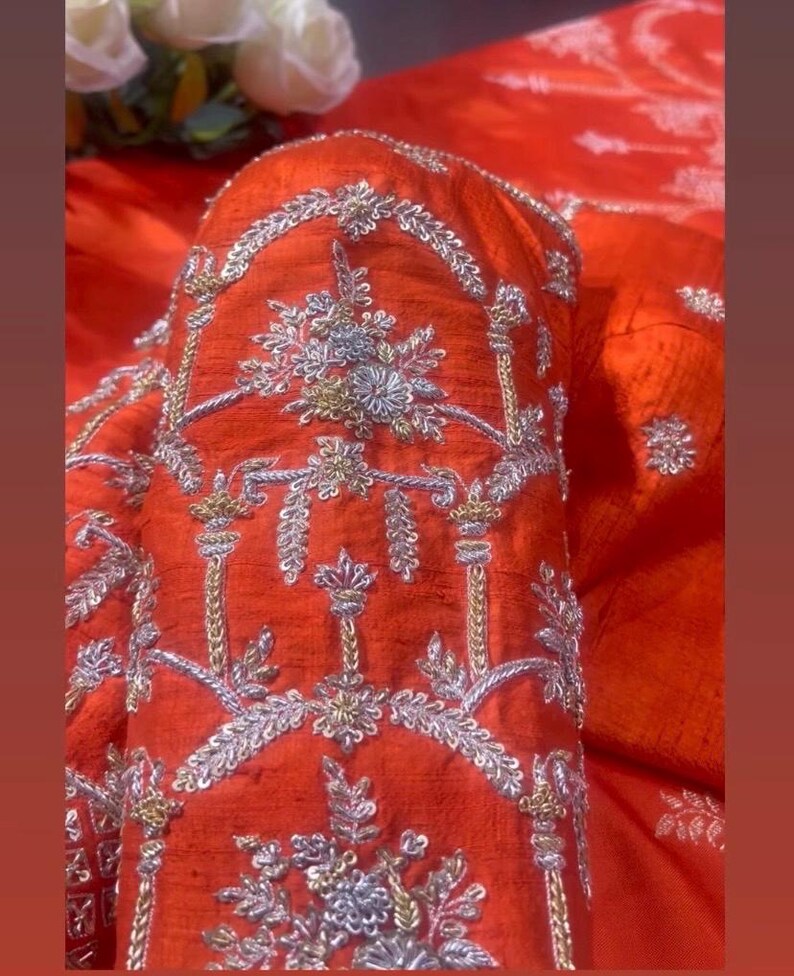 K&ouml;nnte beinhalten: Nahaufnahme eines leuchtend orangefarbenen Kleidungsst&uuml;cks mit aufwendiger Silber- und Goldstickerei. Die detaillierten floralen und geometrischen Muster erzeugen eine luxuri&ouml;se Textur. Der Stoff scheint einen subtilen Glanz zu haben, der den Reichtum des Designs verst&auml;rkt.