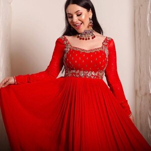 May include: A woman wearing a red, long-sleeved, floor-length gown with silver embroidery and a red and silver jeweled necklace.