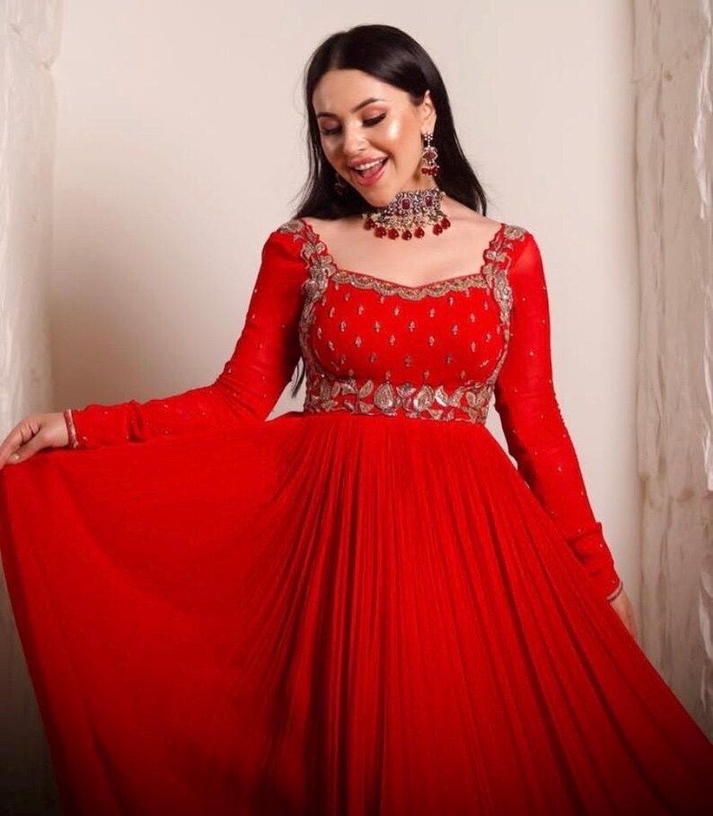 May include: A woman wearing a red, long-sleeved, floor-length gown with silver embroidery and a red and silver jeweled necklace.