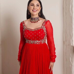 May include: A red floor-length dress with long sleeves and a pleated skirt. The dress has silver embroidery and a red and silver necklace.