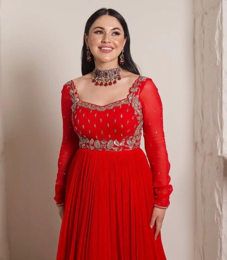 May include: A red floor-length dress with long sleeves and a pleated skirt. The dress has silver embroidery and a red and silver necklace.