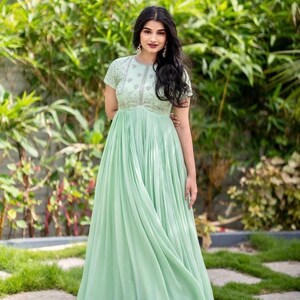 May include: A woman wearing a light green maxi dress with silver embroidery. The dress has a round neckline and short sleeves. The dress is pleated and has a flowing skirt.