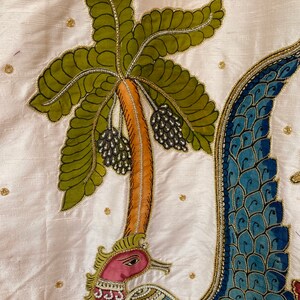 May include: A white fabric with a detailed embroidery of a peacock with a blue and gold tail, a green palm tree with black berries, and a pink flower.