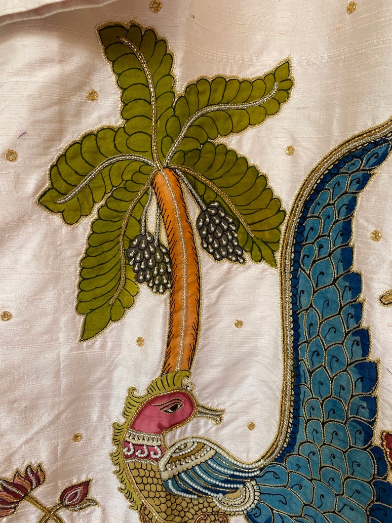 May include: A white fabric with a detailed embroidery of a peacock with a blue and gold tail, a green palm tree with black berries, and a pink flower.