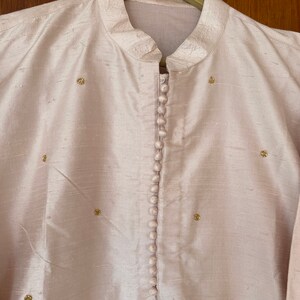 May include: A light pink, long-sleeved shirt with a mandarin collar and a row of buttons down the front. The shirt is decorated with small gold embellishments.