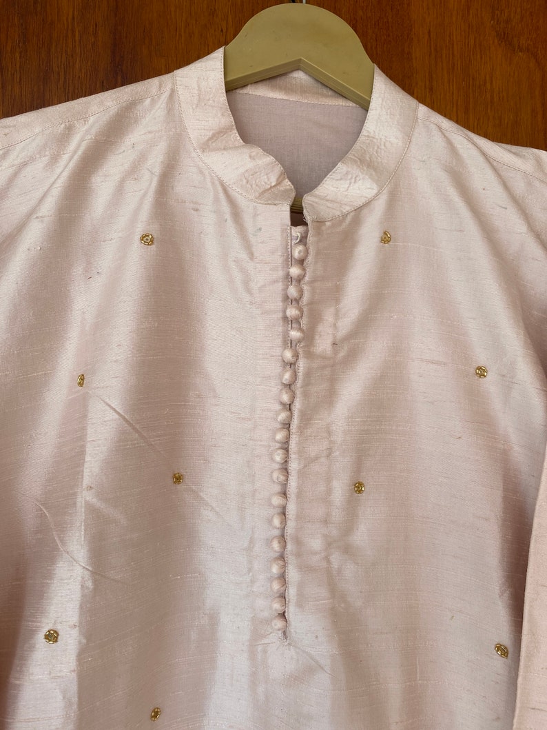 May include: A light pink, long-sleeved shirt with a mandarin collar and a row of buttons down the front. The shirt is decorated with small gold embellishments.