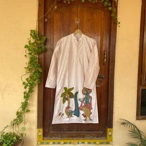 May include: A long-sleeved, light pink kurta with a colorful embroidered design of a figure playing a flute and a palm tree. The kurta is hanging on a hanger in front of a wooden door.