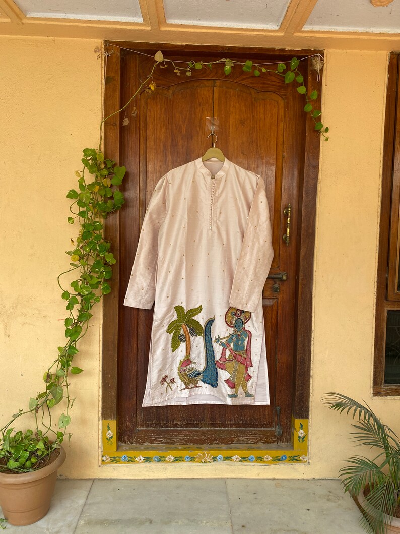 May include: A long-sleeved, light pink kurta with a colorful embroidered design of a figure playing a flute and a palm tree. The kurta is hanging on a hanger in front of a wooden door.