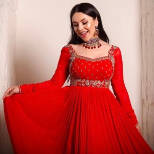 May include: A woman wearing a red, long-sleeved, floor-length gown with silver embroidery and a red jeweled necklace.