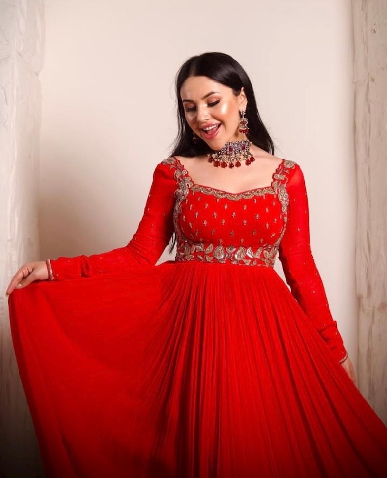 May include: A woman wearing a red, long-sleeved, floor-length gown with silver embroidery and a red jeweled necklace.