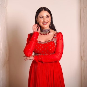 May include: A woman wearing a red dress with long sleeves and a silver and red jeweled necklace. The dress has a square neckline and is embroidered with silver thread. The woman is smiling and looking to the side.