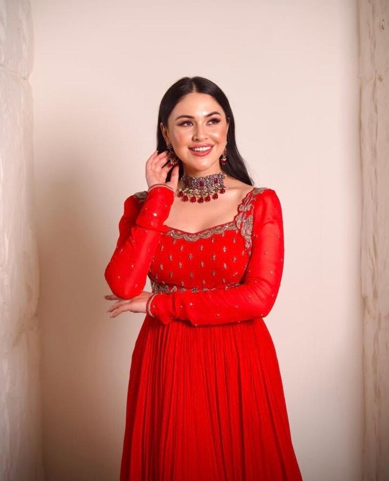 May include: A woman wearing a red dress with long sleeves and a silver and red jeweled necklace. The dress has a square neckline and is embroidered with silver thread. The woman is smiling and looking to the side.