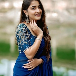 May include: A woman wearing a blue saree with gold embroidery on the sleeves. She is smiling and looking at the camera.