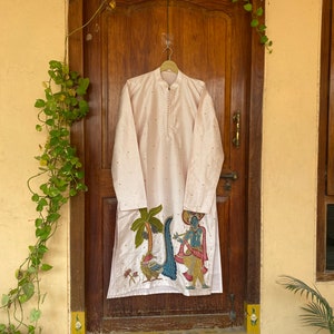 May include: A long, light pink kurta with a white embroidered design of a man playing a flute in front of a palm tree. The kurta has a mandarin collar and a button closure.