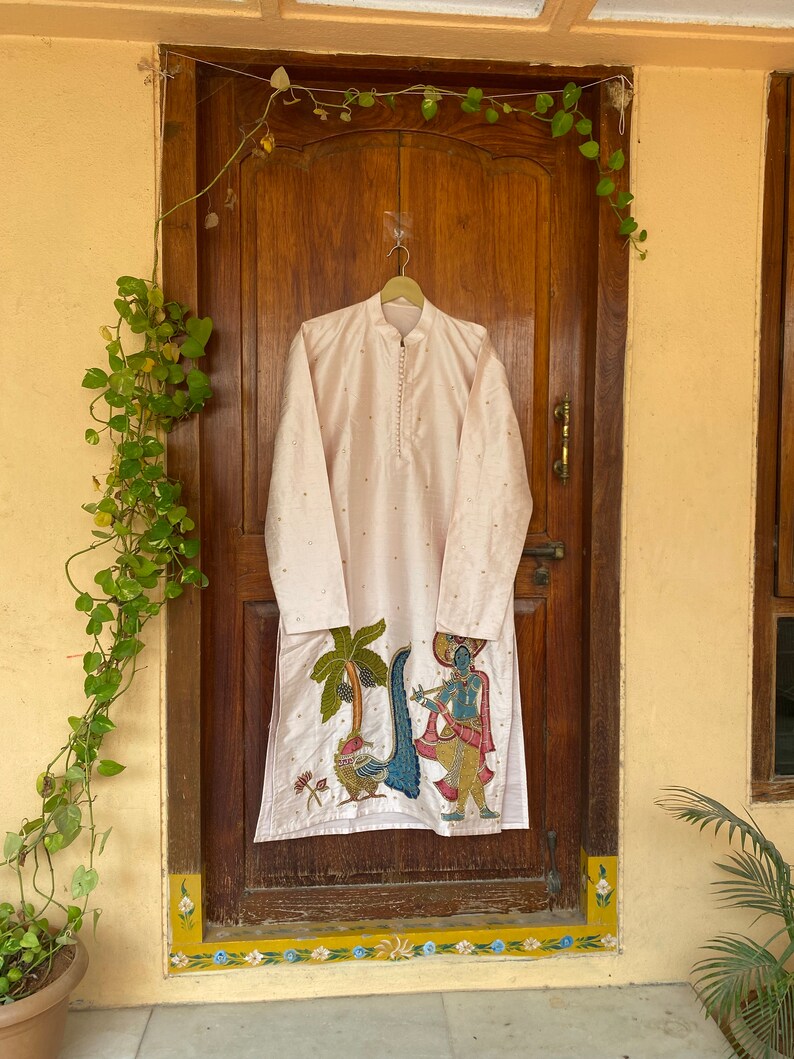 May include: A long, light pink kurta with a white embroidered design of a man playing a flute in front of a palm tree. The kurta has a mandarin collar and a button closure.