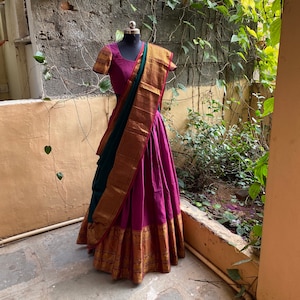 Puede incluir: Un sari indio magenta y verde con ribetes dorados. El sari está drapeado sobre un maniquí y se muestra contra una pared de ladrillo con vegetación.