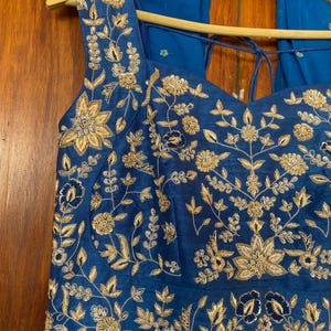 May include: A blue sleeveless dress with gold embroidery and a matching blue scarf. The dress has a sweetheart neckline and a pleated skirt.