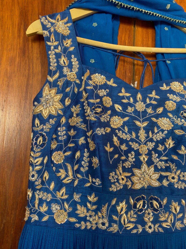 May include: A blue sleeveless dress with gold embroidery and a matching blue scarf. The dress has a sweetheart neckline and a pleated skirt.