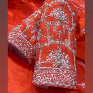 K&ouml;nnte beinhalten: Nahaufnahme eines leuchtend orangefarbenen &Auml;rmels, aufwendig mit Silber- und Goldf&auml;den bestickt. Das Design zeigt florale und geometrische Muster, die eine luxuri&ouml;se und detaillierte &Auml;sthetik schaffen. Die Manschette hat eine detaillierte geometrische Bord&uuml;re.