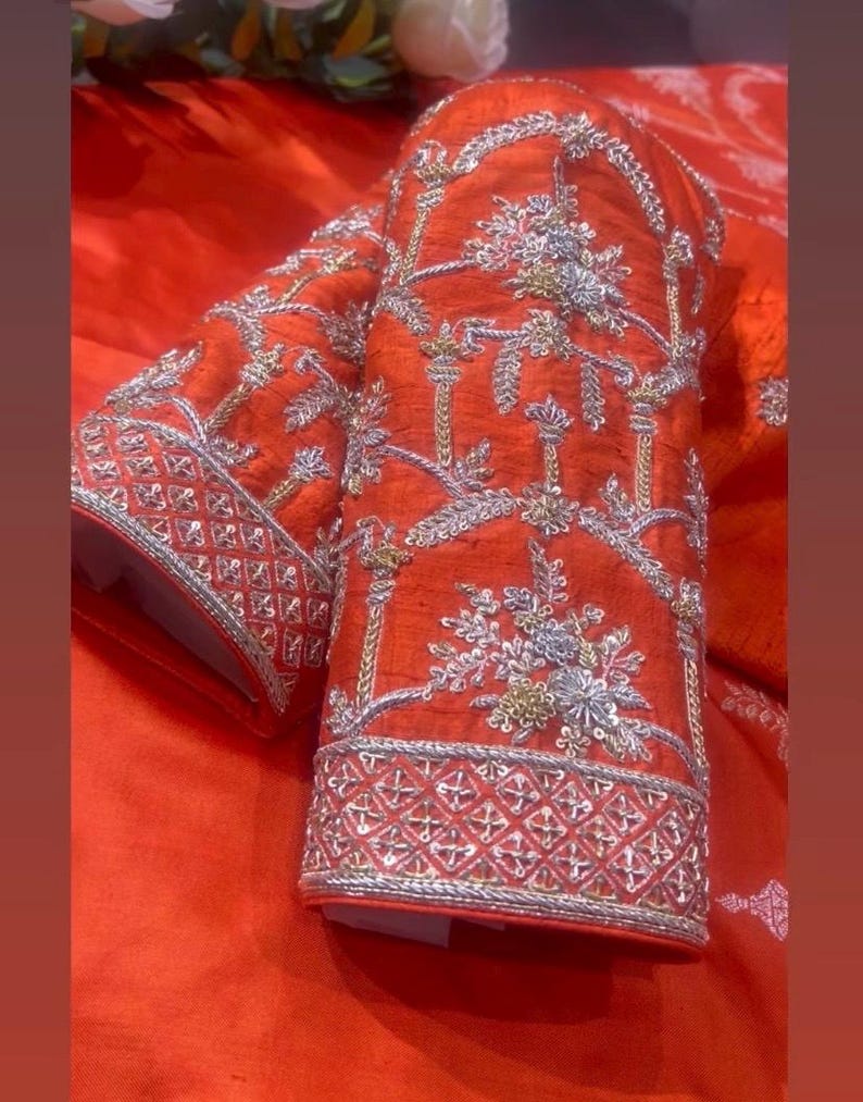 K&ouml;nnte beinhalten: Nahaufnahme eines leuchtend orangefarbenen &Auml;rmels, aufwendig mit Silber- und Goldf&auml;den bestickt. Das Design zeigt florale und geometrische Muster, die eine luxuri&ouml;se und detaillierte &Auml;sthetik schaffen. Die Manschette hat eine detaillierte geometrische Bord&uuml;re.