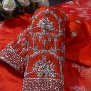 K&ouml;nnte beinhalten: Nahaufnahme eines leuchtend roten &Auml;rmels, aufwendig mit silbernen und goldenen Metallf&auml;den bestickt. Das Design zeigt florale und geometrische Muster, die eine luxuri&ouml;se und detaillierte &Auml;sthetik schaffen. Der &Auml;rmel ist Teil eines gr&ouml;&szlig;eren Kleidungsst&uuml;cks, wahrscheinlich traditioneller oder formeller Kleidung.