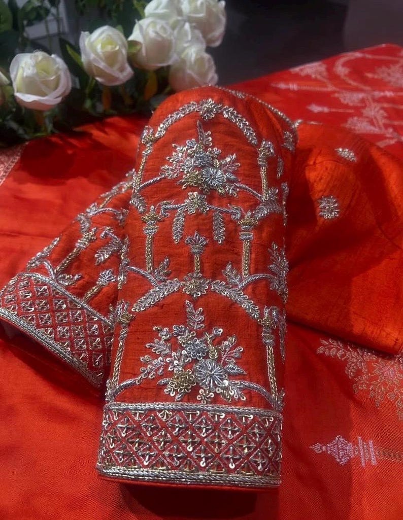 K&ouml;nnte beinhalten: Nahaufnahme eines leuchtend roten &Auml;rmels, aufwendig mit silbernen und goldenen Metallf&auml;den bestickt. Das Design zeigt florale und geometrische Muster, die eine luxuri&ouml;se und detaillierte &Auml;sthetik schaffen. Der &Auml;rmel ist Teil eines gr&ouml;&szlig;eren Kleidungsst&uuml;cks, wahrscheinlich traditioneller oder formeller Kleidung.