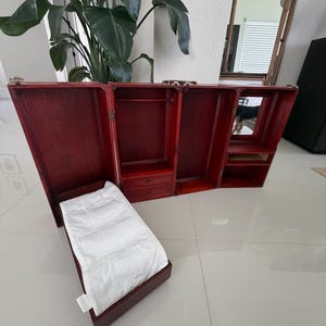 May include: A dark red wooden doll wardrobe trunk that opens into a multi-section display. It features a hanging rod, two small drawers, a mirror, and a separate attached bed section with a white fabric mattress cover and metal latches.