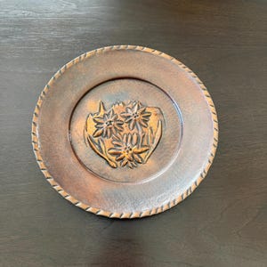 May include: A round copper-toned metal plate featuring a central embossed design of three edelweiss flowers with leaves. The plate has a smooth inner surface and a decorative rope-style textured border along the outer edge.