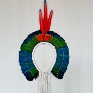 May include: A circular headdress featuring vibrant layers of green, blue, and black feathers arranged in a fan shape. Three long, bright red feathers with blue tips extend upward from the center. The base is a white cord with long white tassels.