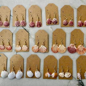 Puede incluir: Una colección de pendientes de conchas marinas exhibidos en etiquetas de cartón marrón. Los pendientes presentan varios tipos y colores de conchas, incluyendo rosa, blanco y naranja, con ganchos dorados. Los pendientes están dispuestos en filas.
