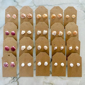 Puede incluir: Una colección de pendientes de concha con tachuelas exhibidos en etiquetas de cartón marrón. Los pendientes presentan varios tipos de conchas en tonos rosa, blanco y beige. Los pendientes están dispuestos en filas, mostrando la variedad.