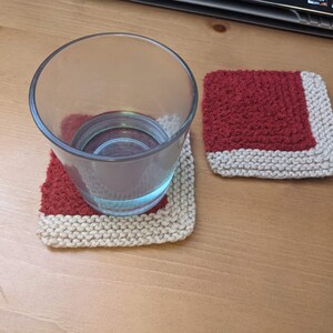 May include: Two knitted coasters with a red and cream color scheme. The coasters are square and have a textured surface. A glass of water sits on one of the coasters.